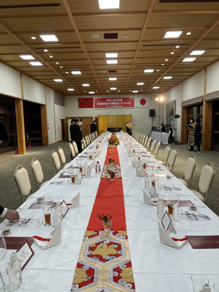 「中華人民共和国 孔鉉佑大使 歓迎レセプション」の準備が整った、栗林公園 商工奨励館（讃岐迎賓館）のホール。広い会場にテーブルと椅子が並ぶ、テーブルには赤いセンタークロスと綺麗な花が飾られている。